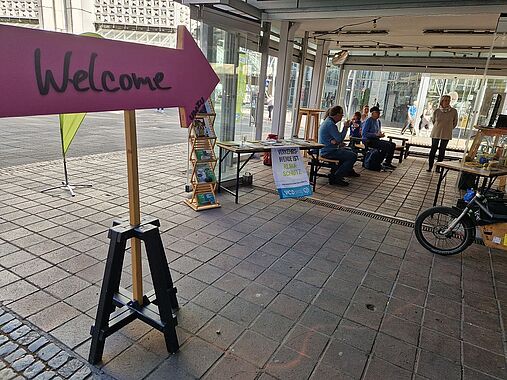 Ein schattiger Mai-Tag mit wenig "Laufkundschaft", das einladende UMZU, Schild "Hereinspaziert" im Vordergrund, Aktive des VCD im Hintergrund