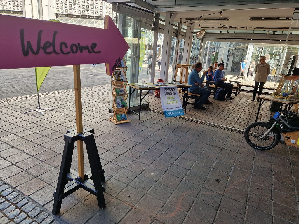 Ein schattiger Mai-Tag mit wenig "Laufkundschaft", das einladende UMZU, Schild "Hereinspaziert" im Vordergrund, Aktive des VCD im Hintergrund